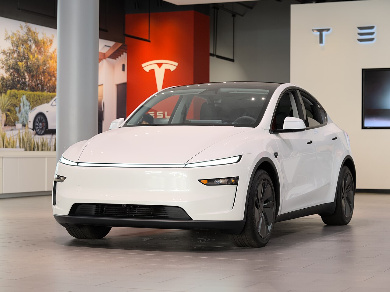 A white Tesla Model Y on a showroom floor, with the Tesla logo on the wall behind it