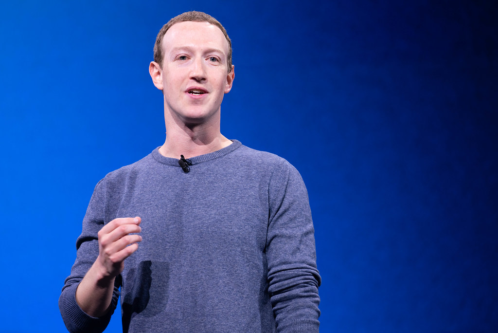 Mark Zuckerberg, in a gray sweater with a clip-on microphone, speaks in front of a deep blue backdrop.