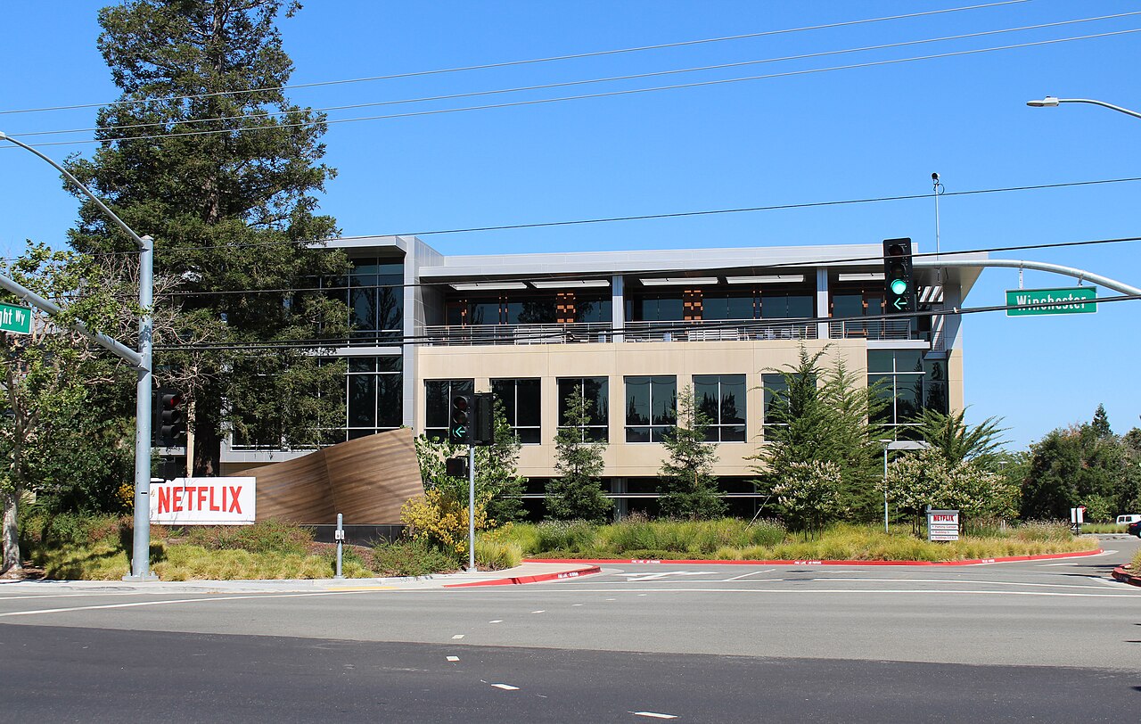 A two-story office building behind a red Netflix sign at a street corner, with trees and a traffic light.