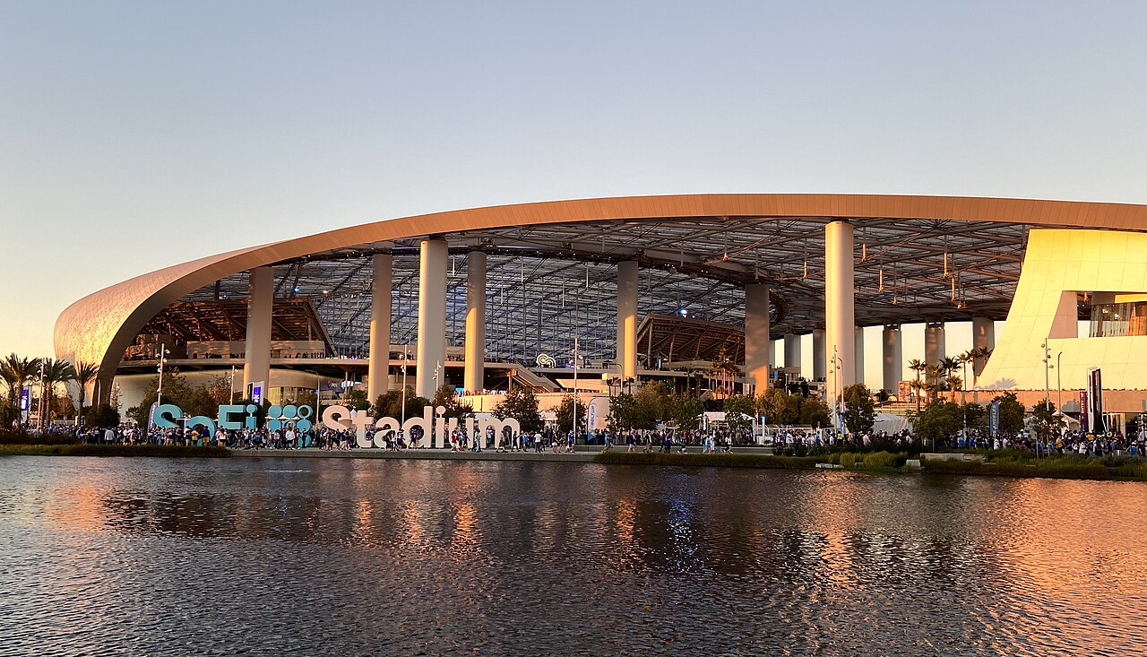 A stadium with a curved canopy roof and white columns, seen across a lake at dusk, with big letters spelling SoFi Stadium in front.